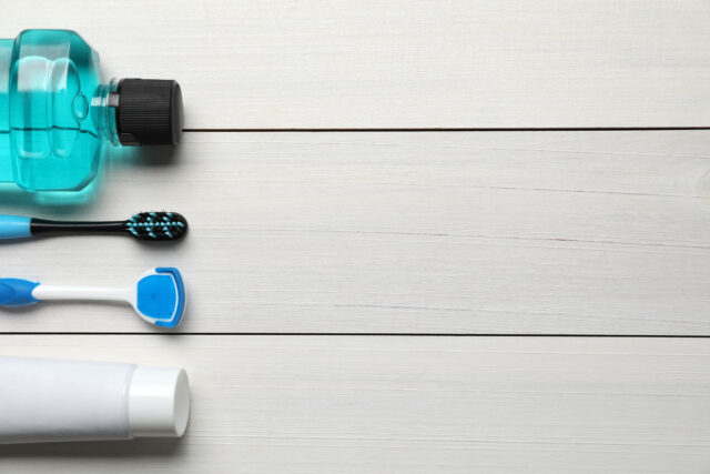 Tongue cleaner, toothbrush, paste and mouthwash on white wooden table.